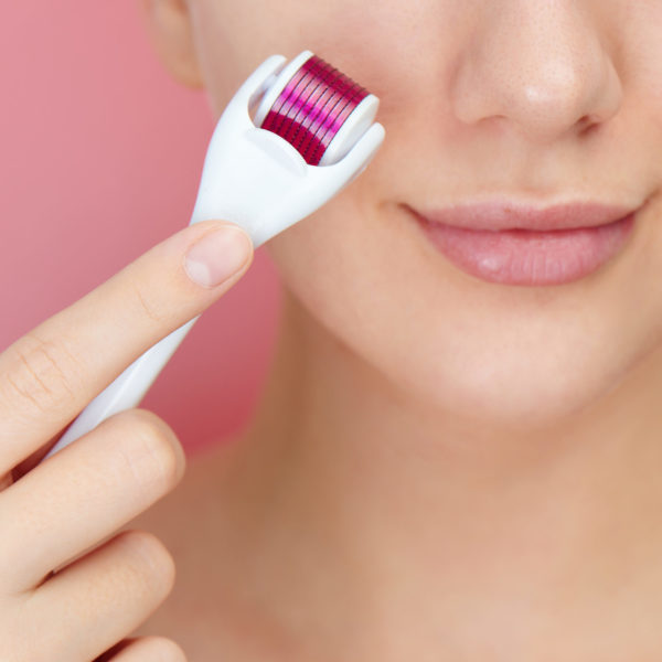 Close-up female face with dermaroller for mesotherapy procedures, skin care at home and in salon. Meso roller with microneedles on pink background